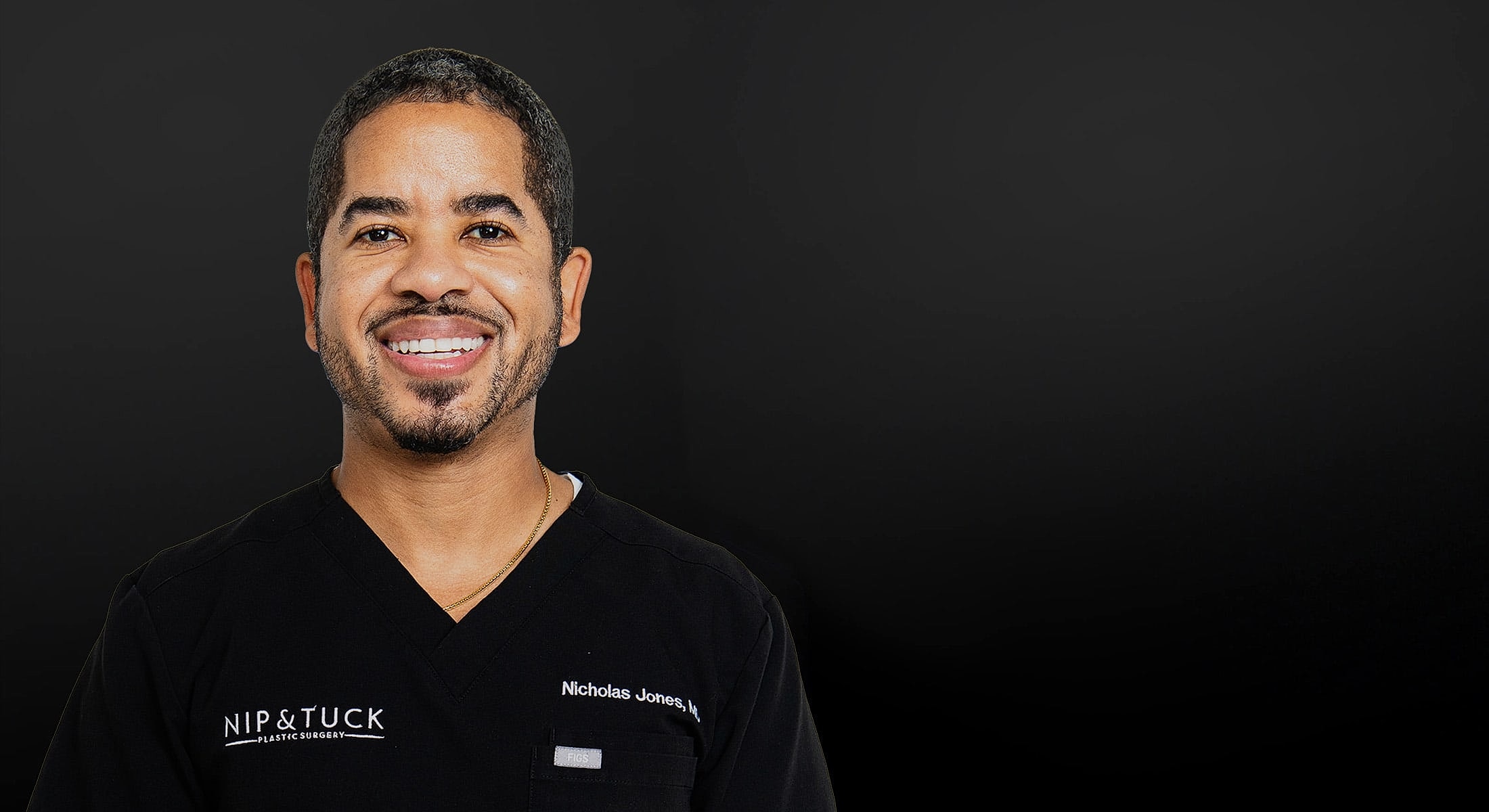 Doctor smiling in scrubs against black background.